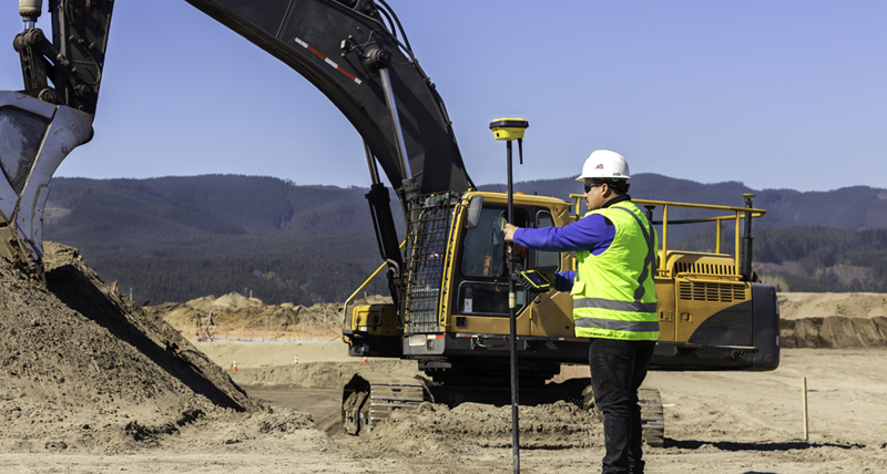 Rakennustyöntekijä keltaisessa suojatakissa mittaa sauvalla Leica iCON GPS 70:n ja Leica iCON CC80 -maastotallentimen kanssa maansiirtotyötä tekevän kaivukoneen edessä.